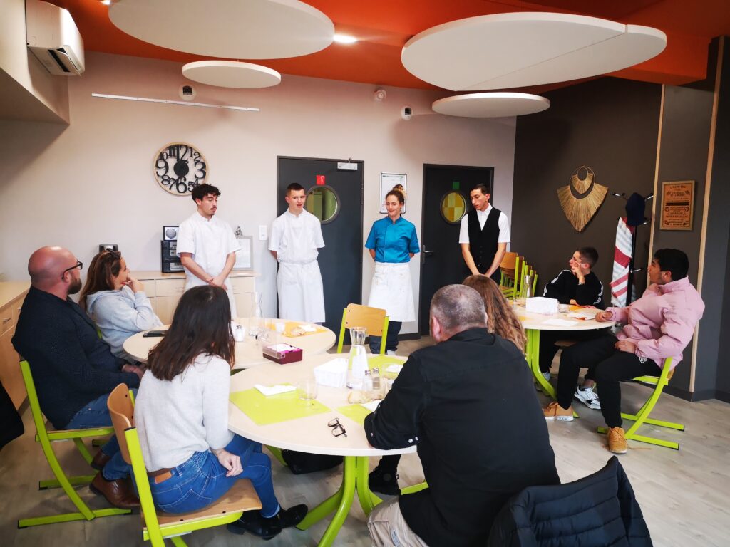 Les "clients" à table et les membres de la brigade, debout, présentent leur menu