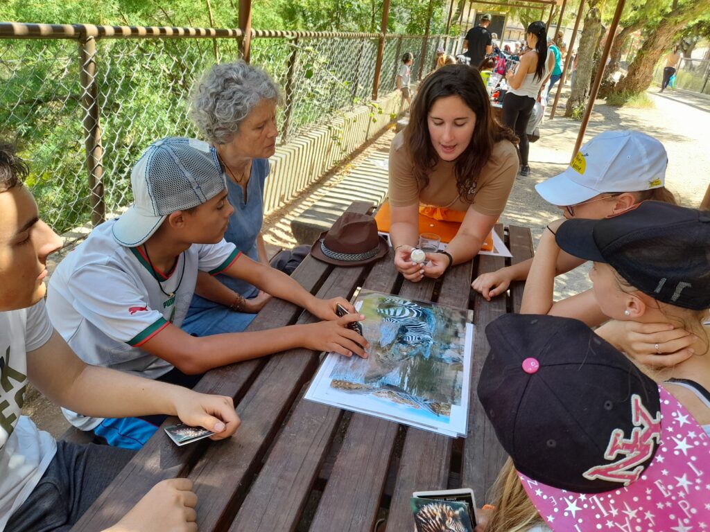 Le groupe, autour d'une table à l'extérieur, écoutent les explications de l'animatrice