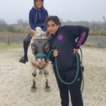 Un jeune accompagné est sur un poney qui est tenu par la bride par une monitrice