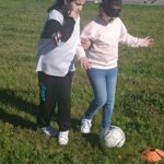 Une jeune fille les yeux bandés et le ballon de foot aux pieds, accompagnée d'une autre jeune fille qui la guide, sur un terrain de foot