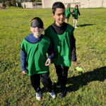 Un garçon les yeux bandés guidé par un autre garçon sur un terrain de foot et à l'arrière plan, un binôme de jeunes filles