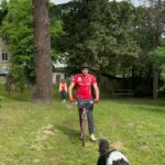 Un jeune homme sur un vélo avec un casque, tiré par un chier en laisse qui court. En arrière plan une personne regarde