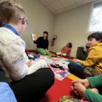 Assise au sol, une animatrice lit un livre aux enfants et leur accompagnant tous assis en demi cercle sur un tapis