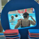 Jeu de ballon dans l'eau. De dos, une jeune femme garde une espèce de cage en mousse bleue. En arrière plan 4 personnes dans l'eau dont une qui semble garder une autre cage rouge en face