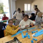 11 personnes (enfants et adultes) autour des tables confectionnent les éléments pour le corso fleuri