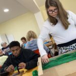 6 personnes (enfants et adultes) autour des tables confectionnent les éléments pour le corso fleuri