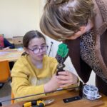 Une jeune fille confectionne une fleur avec le soutien d'une bénévole de l'association "Char fleuri". En arrière plan, une bénévole de l'association "Char fleuri" travaille également