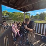L'animateur regarde dans le grand objectif d'un appareil photo. A ces côtés 4 jeunes le regardent