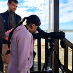 Une jeune regarde dans le grand objectif d'un appareil photo. En arrière plan l'animateur