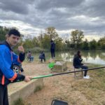 AU premier plan un jeune fait un pouce à l'objectif avec sa canne à pêche dans la main et en arrière plan des jeunes pêchent près du plan d'eau