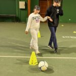 Dans le gymnase, une jeune fille guide un jeune garçon avec les yeux bandés qui doit faire un parcours avec un ballon de football.