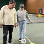 Dans le gymnase, une jeune fille tape dans un ballon les yeux bandés. Auprès d'elle, un adulte la guide.