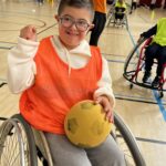 Sur le parquet du gymnase, un jeune garçon sur un fauteuil roulant avec un ballon en mousse sur les genoux. A l'arrière plan d'autres jeunes en fauteuils roulants.