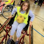 Sur le parquet du gymnase, une jeune fille sur un fauteuil roulant avec un ballon en mousse sur les genoux. A l'arrière plan, d'autres jeunes en fauteuils roulants.