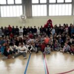 Sur le parquet du gymnase, photo de groupe de l'ensemble des participants avec la mascotte des JO de Paris